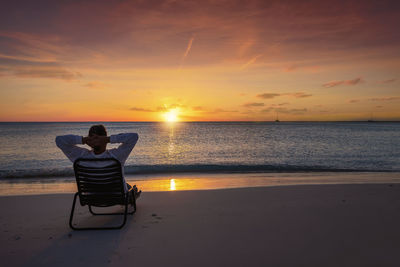 Rear view of woman sitting on beach against sky during sunset