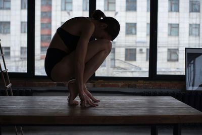 Side view of young woman exercising on table against window