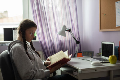 A teenage girl with glasses reads a book at her desk at home.