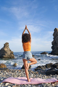 Full body back view of anonymous barefoot female practicing vrikshasana posture with raised arms during yoga training on stony coast near sea