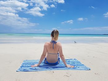 Rear view of mid adult woman in bikini sitting on blanket at beach against blue sky