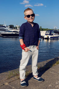 Baby boy blond in blue shirt, light trousers and sun glasses walking on promenade  water with yachts