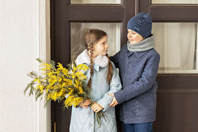 A couple of a boy and a girl of schoolchildren in spring coats with a bouquet of mimosa in  hands 