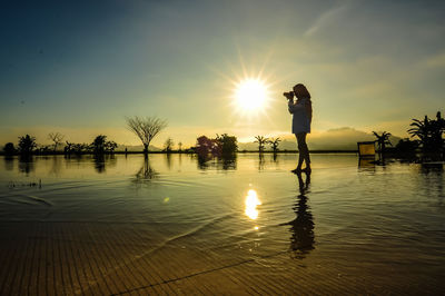 Silhouette woman photographing by lake against sky during sunset