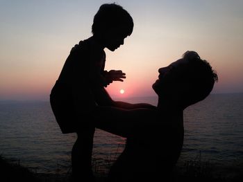 Side view of silhouette man with son standing at beach against sky during sunset