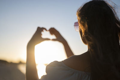 A girl in a dress on the sea at sunset shows a heart with her hands