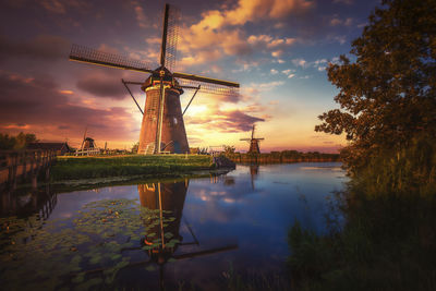 Traditional windmill by lake against sky during sunset