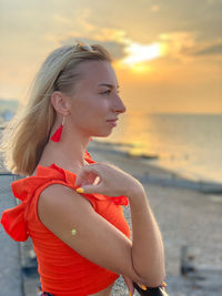 Young woman looking away while standing on beach against sky during sunset