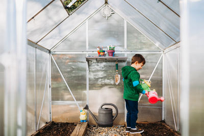 Young boy holding watering cans in backyard greenhouse in springtime