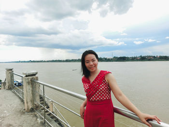 Portrait of smiling young woman standing on railing against sky