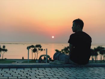 Rear view of man sitting by sea against sky during sunset