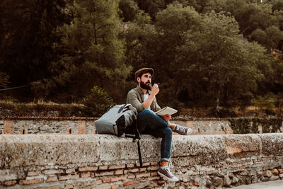 Man with backpack holding pencil and book while sitting outdoors