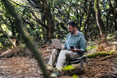 Full length of man using laptop while holding mobile phone sitting in forest