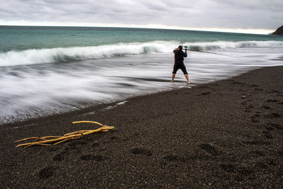 Rear view of man fishing in sea against cloudy sky