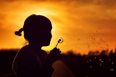 Side view of silhouette girl blowing dandelion seed against sky during sunset