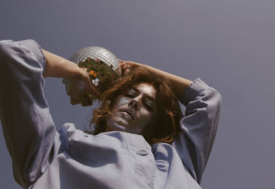 Low angle portrait of woman with glitter on face holding disco ball against clear sky