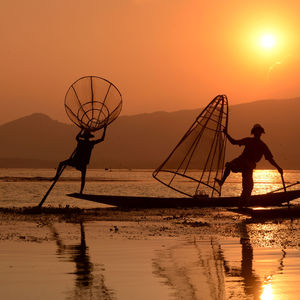 Silhouette fishermen balancing on boat in lake against sky at sunset