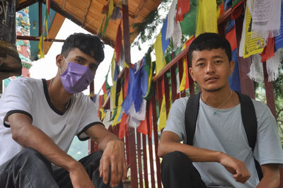 Two male friends looking at camera with sitting on stairs of buddhist temple during covid-19 