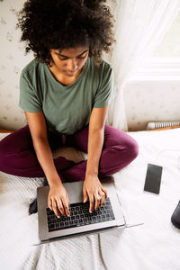 High angle view of young woman with curly hair using laptop while sitting on bed at home