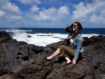 Woman sitting on rock at beach against sky