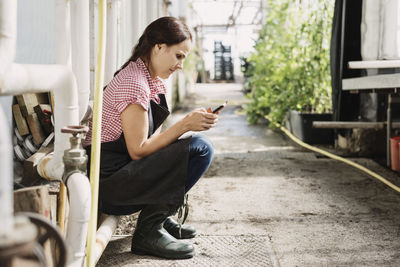 Female gardener text messaging on mobile phone while sitting in greenhouse