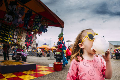 Cute girl wearing sunglasses eating cotton candy while standing against sky at amusement park