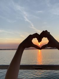 Person hand holding heart shape against sea during sunset