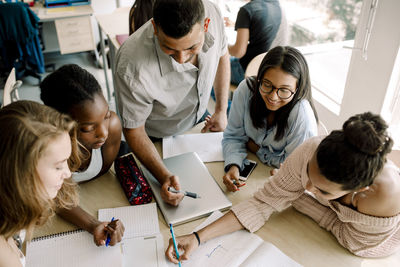 High angle view of teenagers studying while professor standing by table in classroom