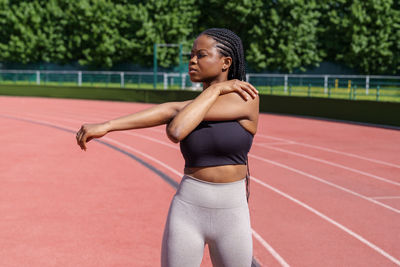 Black young woman with long braids stretches hands and shoulders standing on red track on stadium