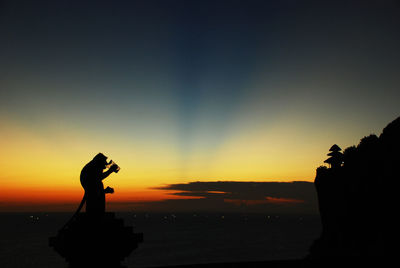 Silhouette a monkey drinking a cup mineral water on rock by sea against sky during sunset