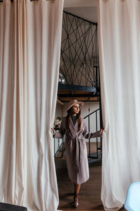 A girl in a beige hat and a beige coat stands indoors and pushes the white curtain with her hands
