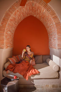 Full length of woman sitting on sofa against orange wall