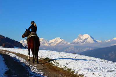 Rear view of woman riding horse on snow covered mountain against clear sky