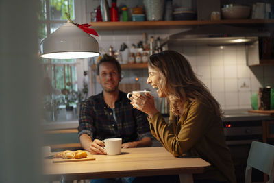 Cheerful woman holding coffee cup sitting with boyfriend at dining table in kitchen