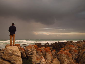 Rear view of man standing on rock formation against cloudy sky