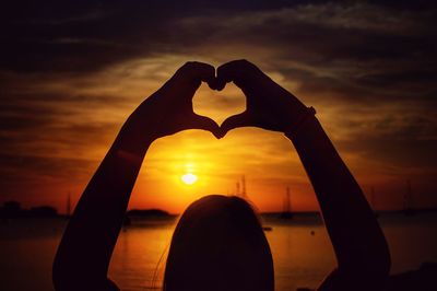 Silhouette woman at lakeshore making heart shape against cloudy sky during sunset