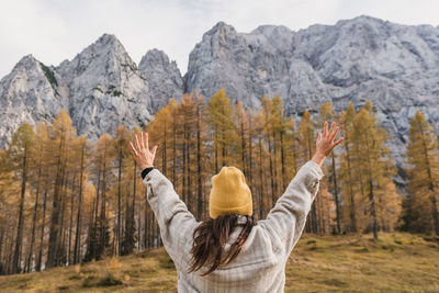 Rear view of young woman with raised arms standing under beautiful mountains in autumn