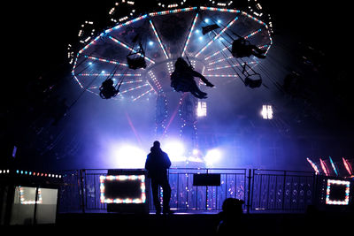 Silhouette man standing against illuminated chain swing ride at night