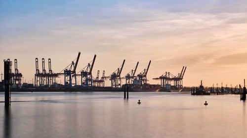 Cranes at harbor against sky during sunset