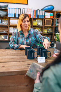 Female customer holding her purse to pay while happy cashier offering discount mugs on local store