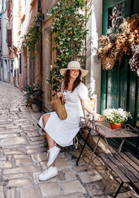 Portrait of a beautiful young woman in white dress sitting on a chair in old town street.