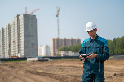 A man in a helmet and overalls controls a drone at a construction site. the builder carries out