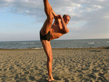 Portrait of shirtless mature man exercising at beach against sky during sunset