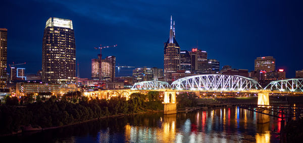 Illuminated bridge over river by buildings against sky at night