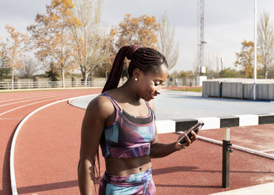 Smiling sportswoman with braided hair using smart phone on track during sunny day