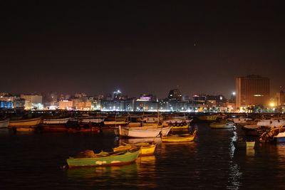 Boats moored on river by illuminated city against sky at night