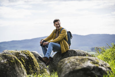 Happy mature man with backpack sitting on rock in front of sky