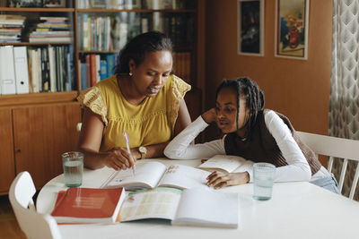 Mother explaining daughter over book at table in living room