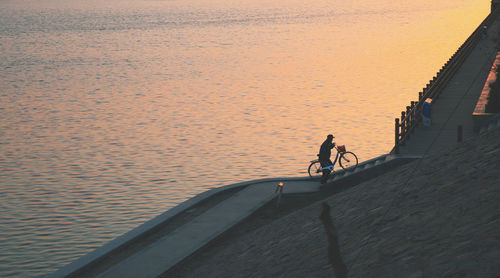 Man riding bicycle by sea against sky during sunset