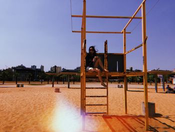 Woman sitting on outdoor play equipment against clear blue sky